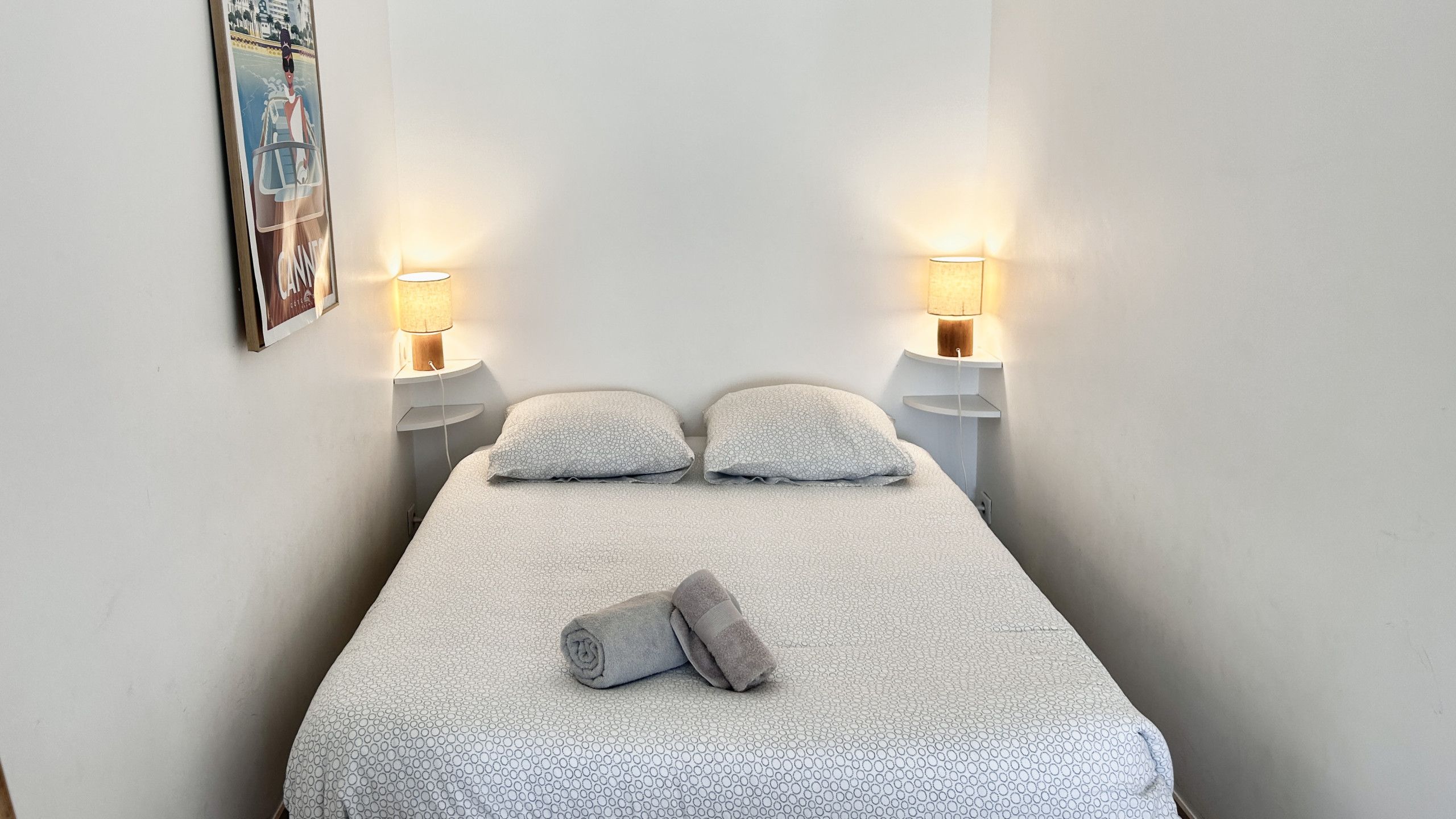 Ein Einzelbett mit einem gemusterten Bettbezug, zwei Kissen, zwei Nachttischlampen und ein zusammengerolltes Handtuch.
