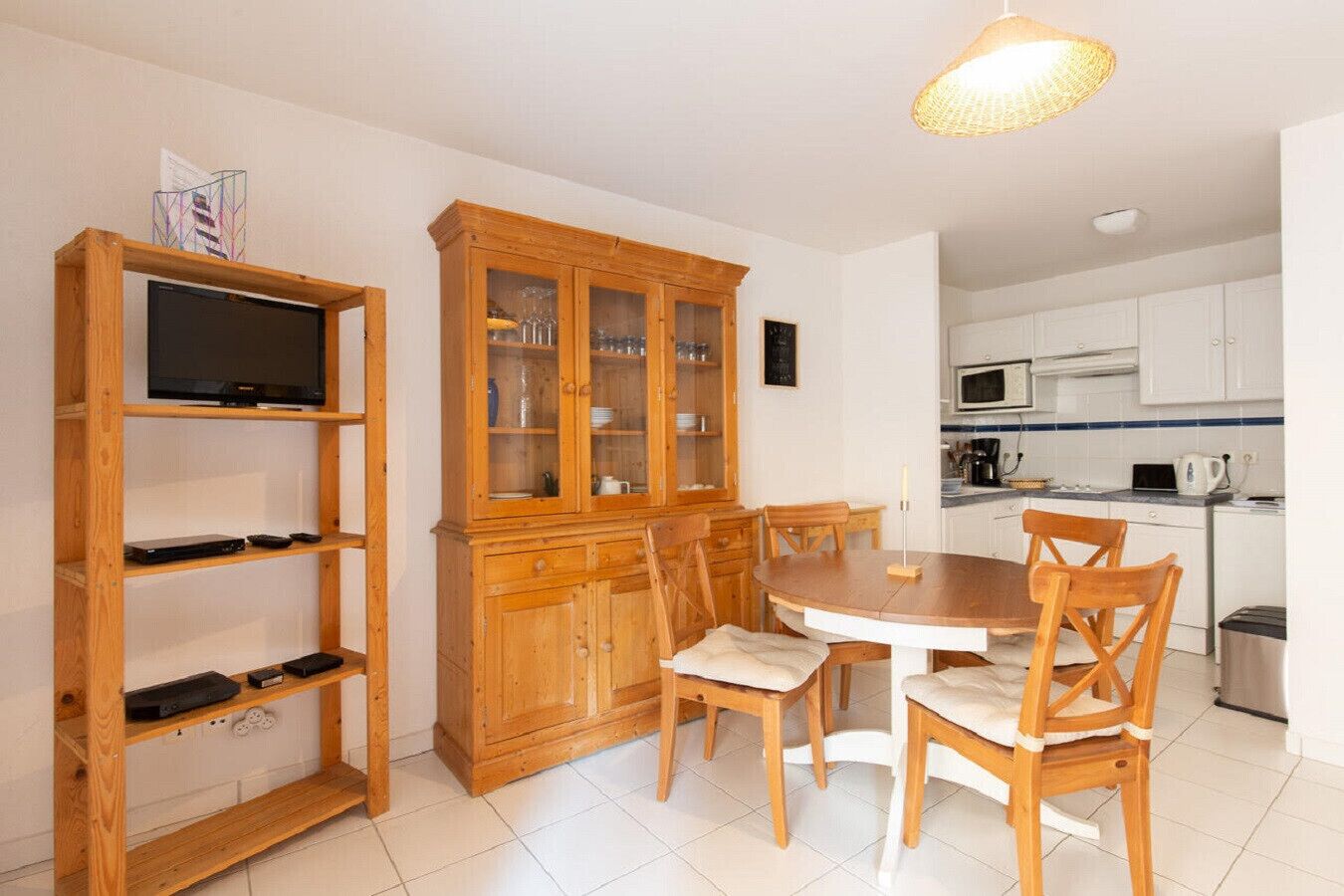Eine kleine Küche mit Esstisch und Stühlen, einem Holzschrank mit Glastüren und einem Fernseher auf einem Regal.