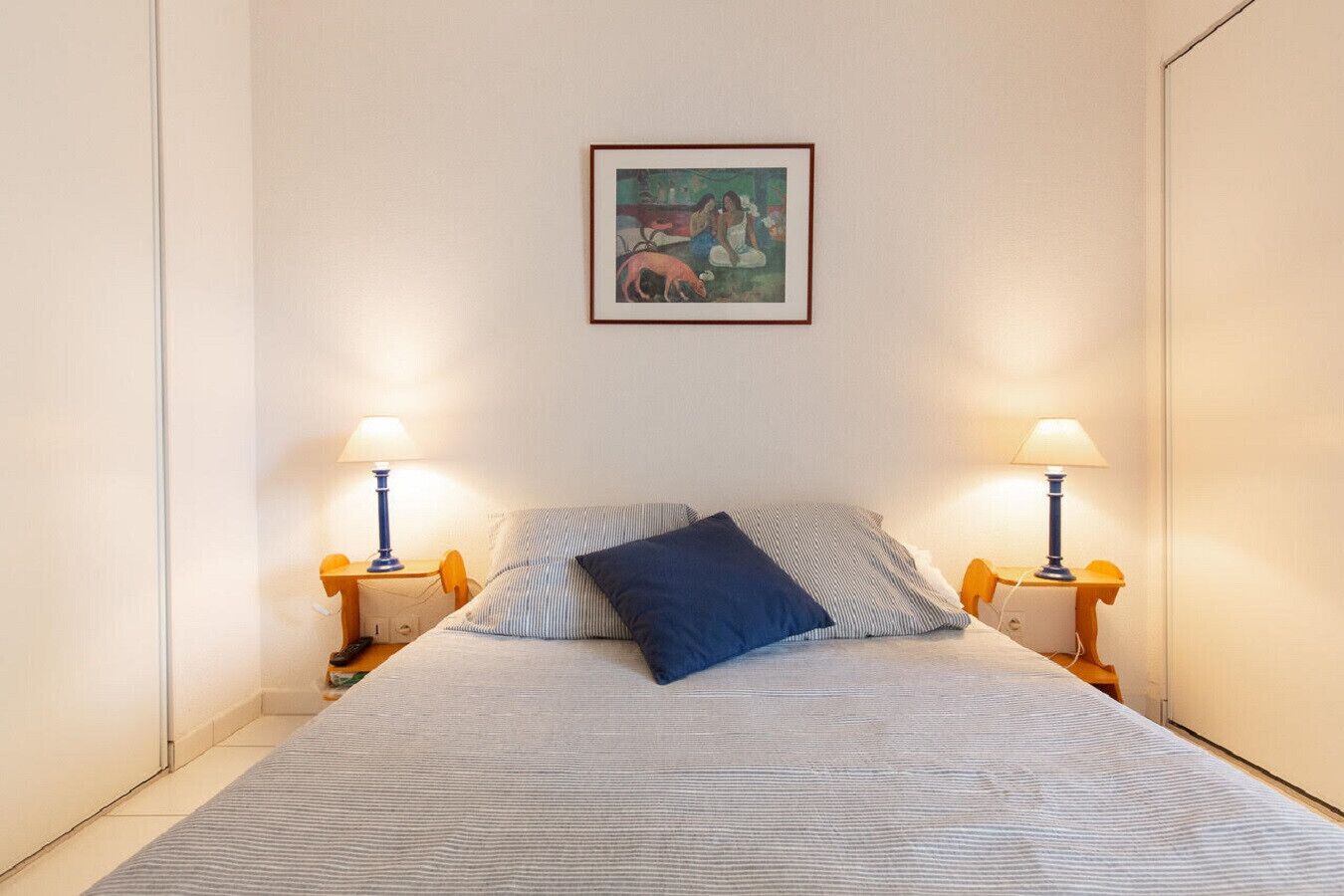A bedroom with a bed, two bedside lamps, a painting on the wall, and a small table with a lamp.