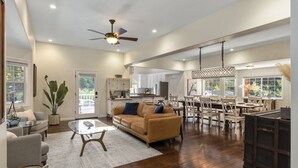 A spacious living area with a sofa, coffee table, and dining table. There is a ceiling fan, a kitchen in the background, and large windows.