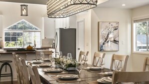 A dining room with a large wooden table set for six, surrounded by wooden chairs. A chandelier hangs above the table, and a large window provides natural light.