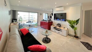 A modern living room with a sofa, a glass coffee table, a red kitchen island, a flat-screen TV, and a large window offering a view of the outside.