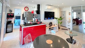 A modern kitchen with a red island, a dining area with a glass table, and a living space with a TV and plant.