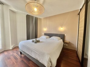 A hotel room with a large bed, two bedside lamps, a window with curtains, and a wooden floor. - Carre d'or duplex (Cannes)