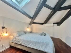A bedroom with a sloped ceiling, exposed wooden beams, a bed with patterned bedding, two wall-mounted lamps, and a radiator. - Carre d'or duplex (Cannes)