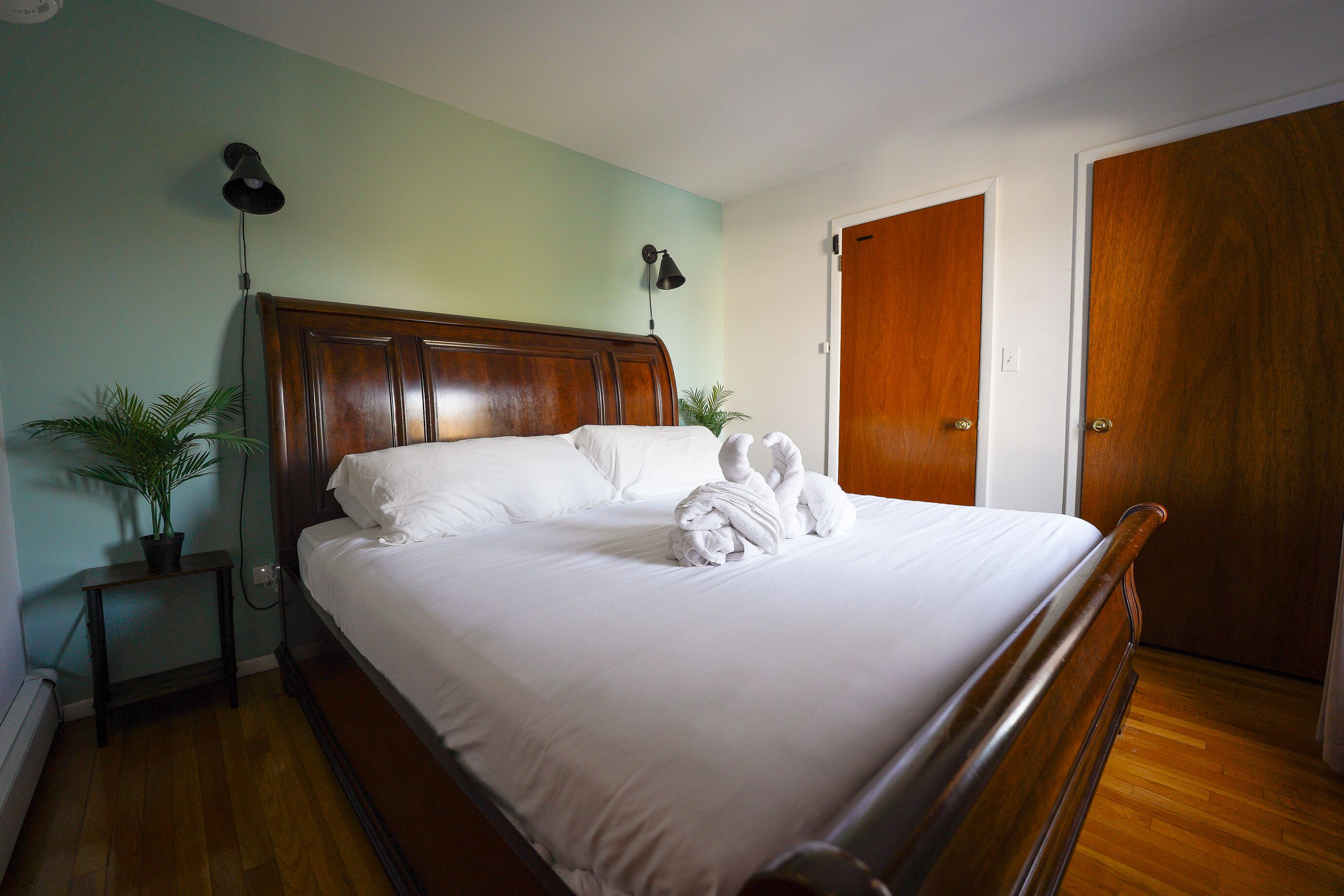 A neatly made bed with white linens and towels, a wooden headboard, a small bedside table with a lamp, and a potted plant.