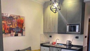 A compact living space with a grey sofa, a small round table, a washing machine, a microwave, and a hanging light fixture.