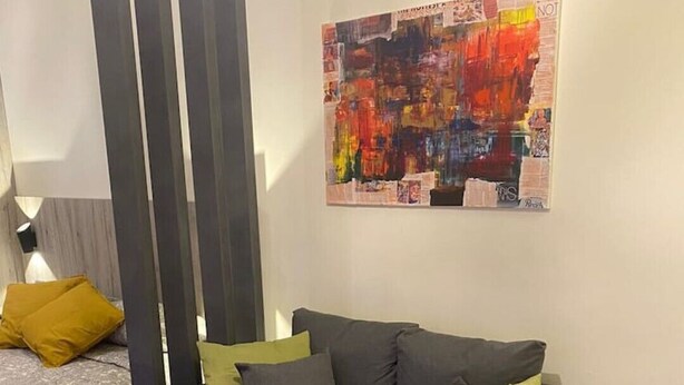 Ein modernes Wohnzimmer mit grauem Sofa, grünen und gelben Kissen, einem farbenfrohen abstrakten Gemälde an der Wand und einem grauen Hochflor Teppich.