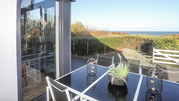 A modern outdoor dining area with a glass table and chairs, a potted plant, and a view of the sea.