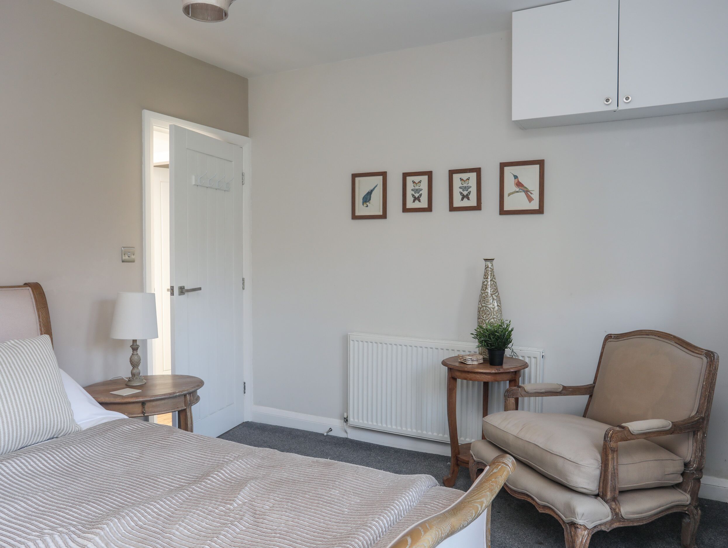 A bedroom with a bed, a chair, a nightstand, a lamp, a radiator, and framed pictures on the wall.