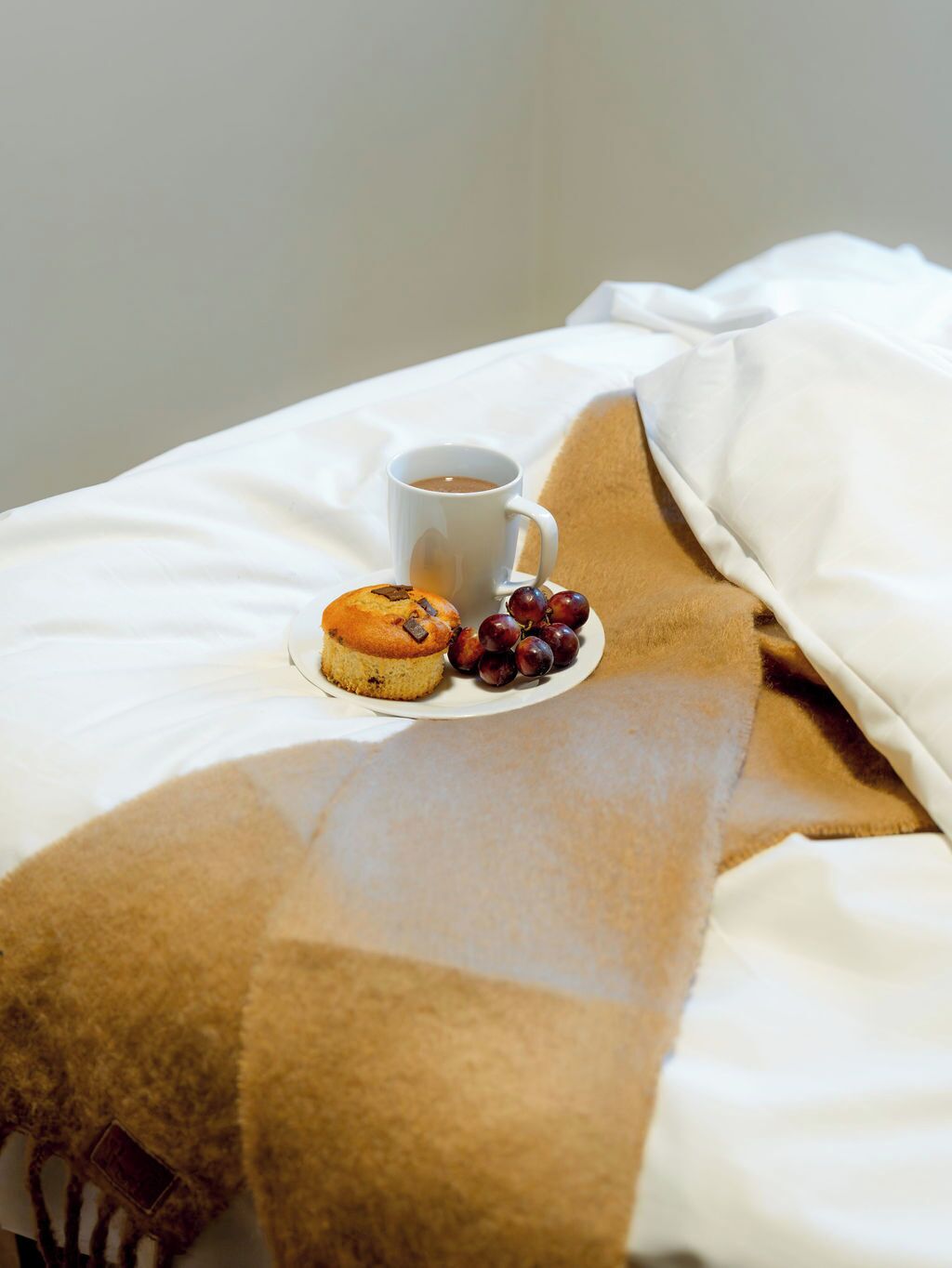 A bed with a white sheet, a brown blanket, a plate with a muffin and grapes, and a cup of coffee.