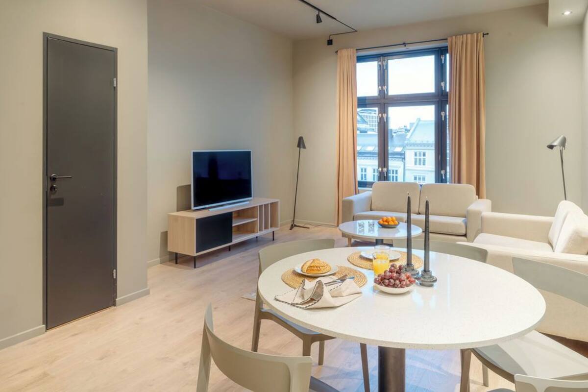 A modern living room with a flat-screen TV, a dining table with breakfast items, a sofa, and a large window with curtains.