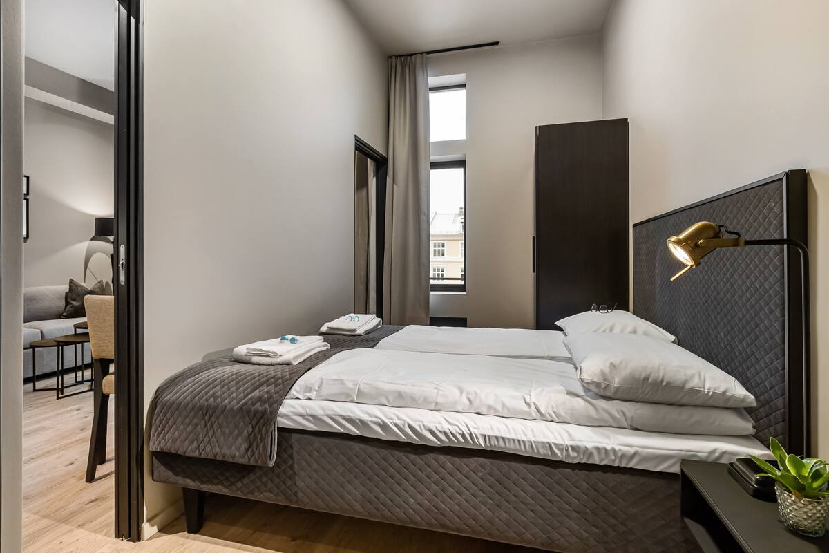 A modern hotel room with a large bed, a bedside table with a lamp, a small plant, and a view of the city through a window.