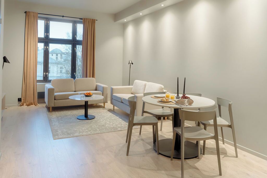 A modern living room with a sofa, a coffee table, and a dining table with chairs. There is a large window with curtains, a floor lamp, and a view of buildings outside.