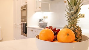 Une cuisine avec des armoires blanches, un plan de travail blanc et un saladier contenant des oranges et un ananas.