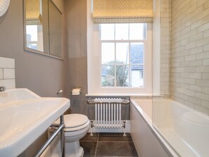 A compact bathroom with a white sink, toilet, and bathtub. A window with a patterned valance allows natural light, and a radiator is visible.