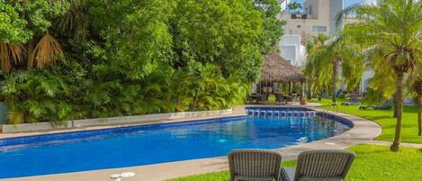 A swimming pool with lounge chairs, surrounded by lush greenery and a thatched-roof structure.