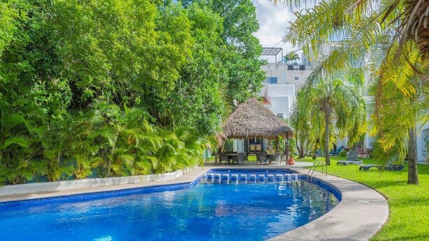 A swimming pool with a thatched-roof structure, surrounded by lush greenery and palm trees. - Playacar Townhome: Pool, Patio & Beach Walk (Playa del Carmen)