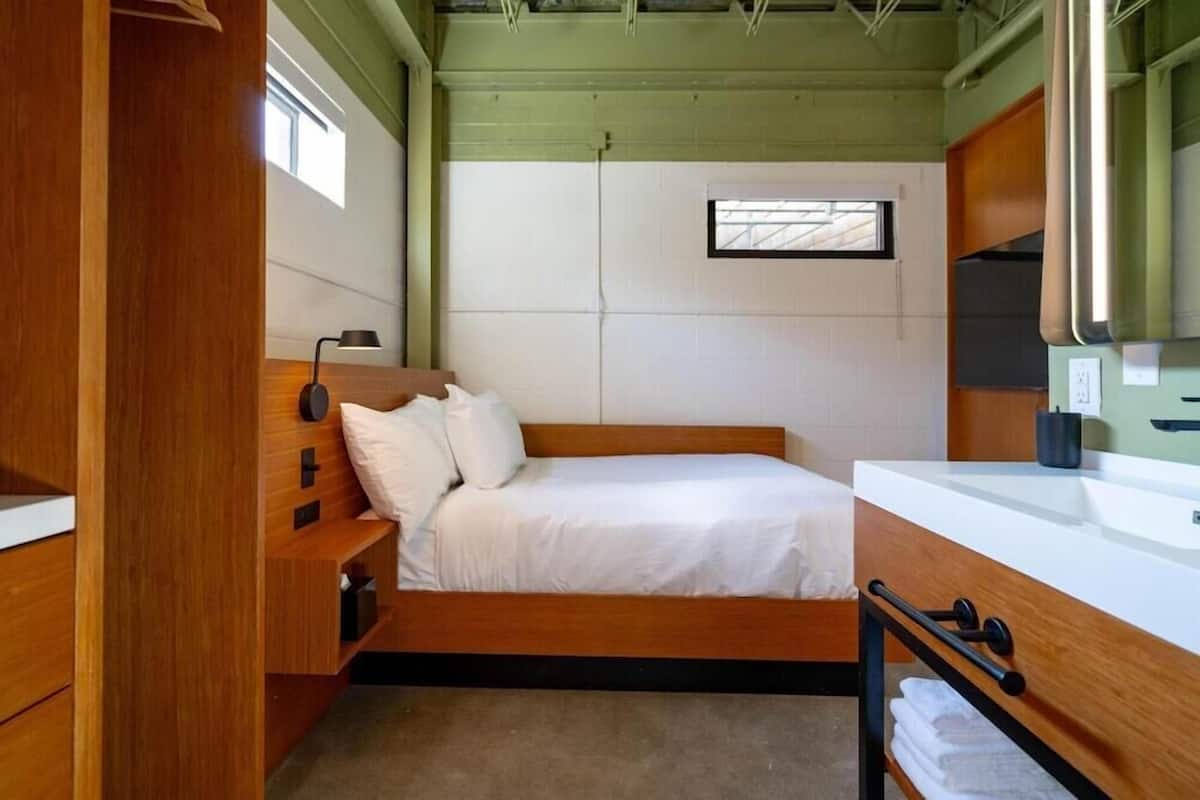 A compact bedroom with a single bed, a bedside table, a small window, and a bathroom with a mirror and sink.