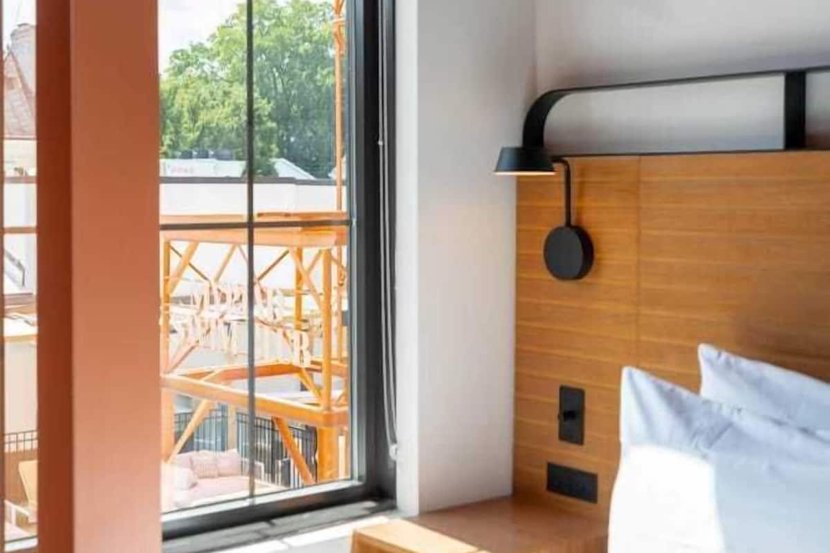 A modern hotel room with a large window, a wooden headboard, a bedside table, and a bed with white bedding.