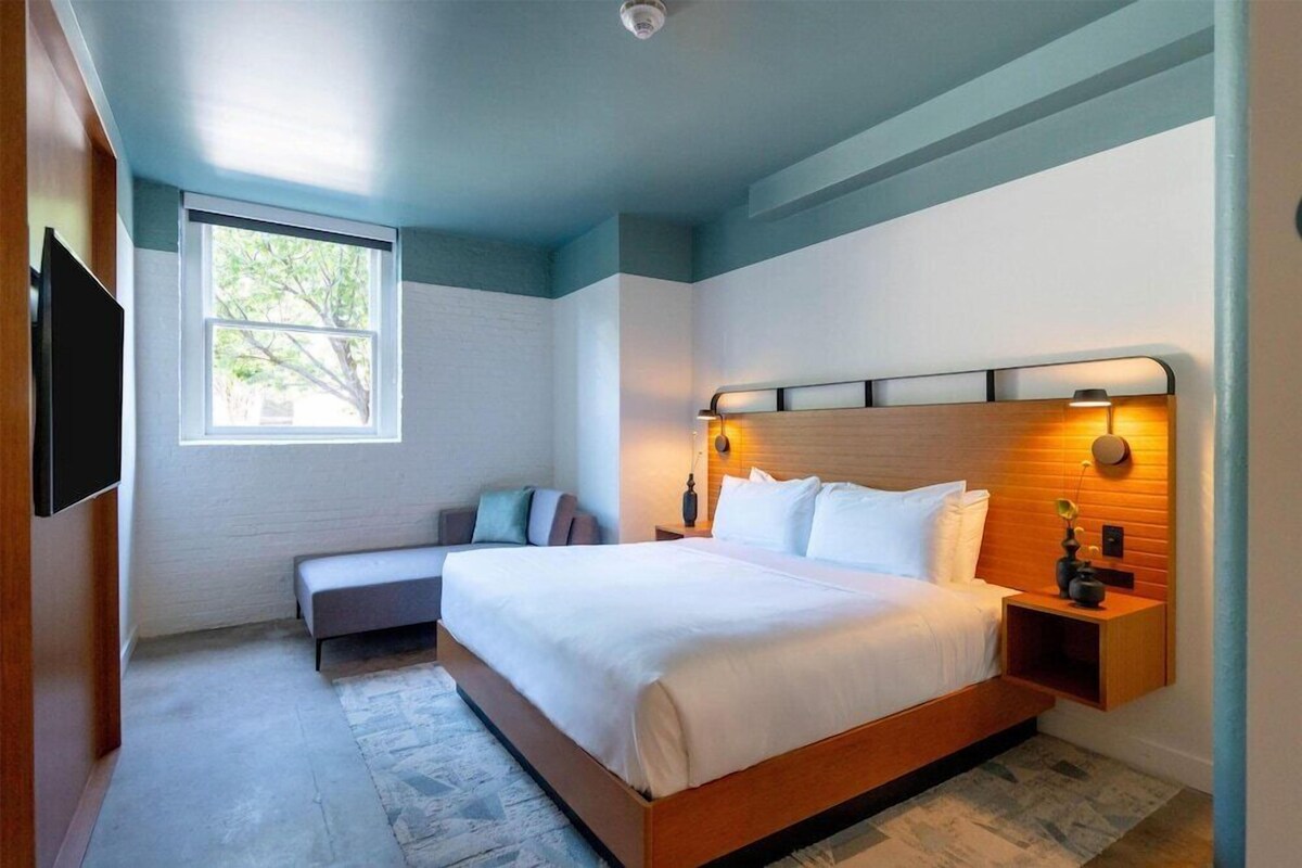 A modern hotel room with a large bed, a bench, a TV, and a window with a view of trees.