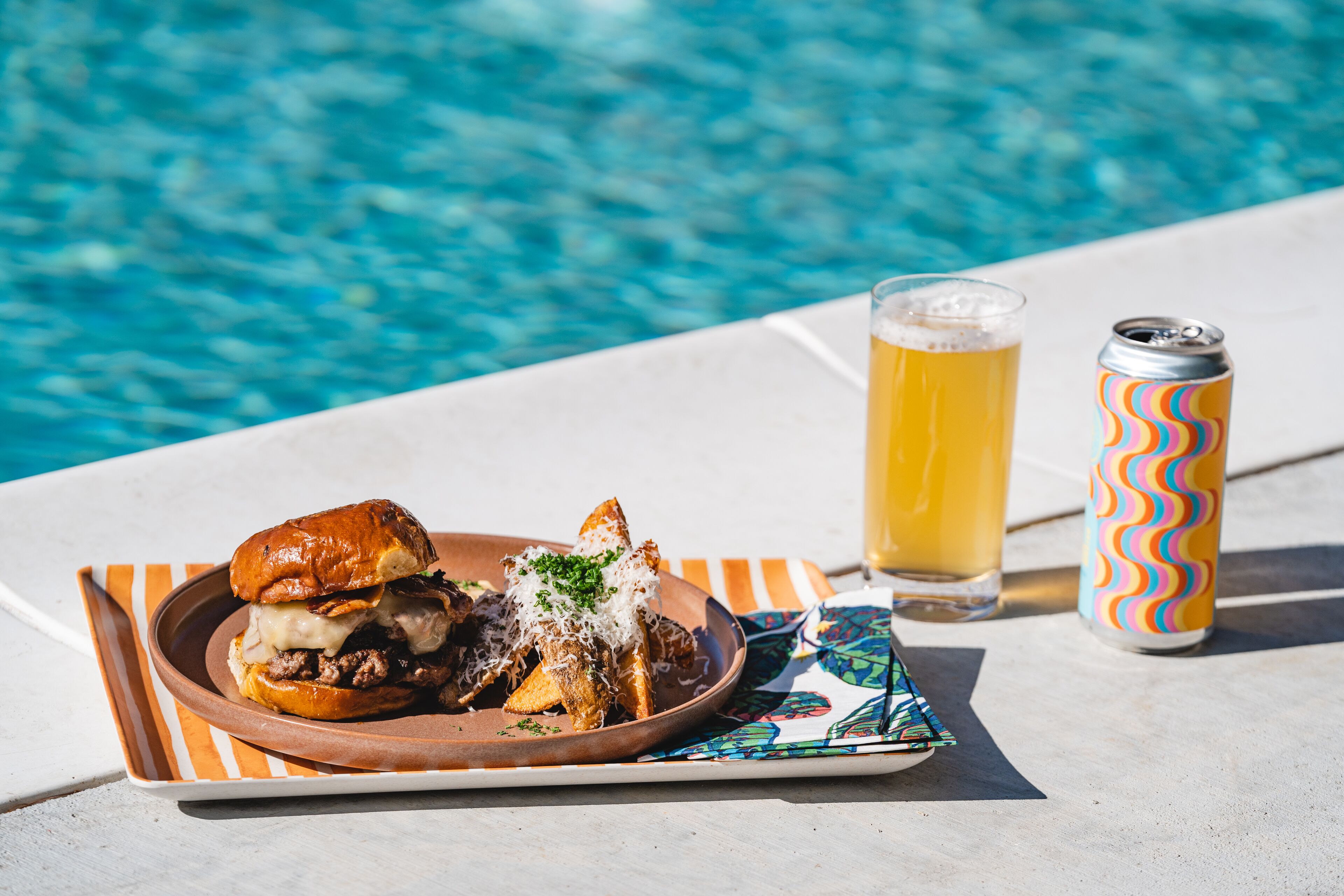 Un plato con una hamburguesa y papas fritas, una jarra de cerveza y una lata al lado de una piscina.