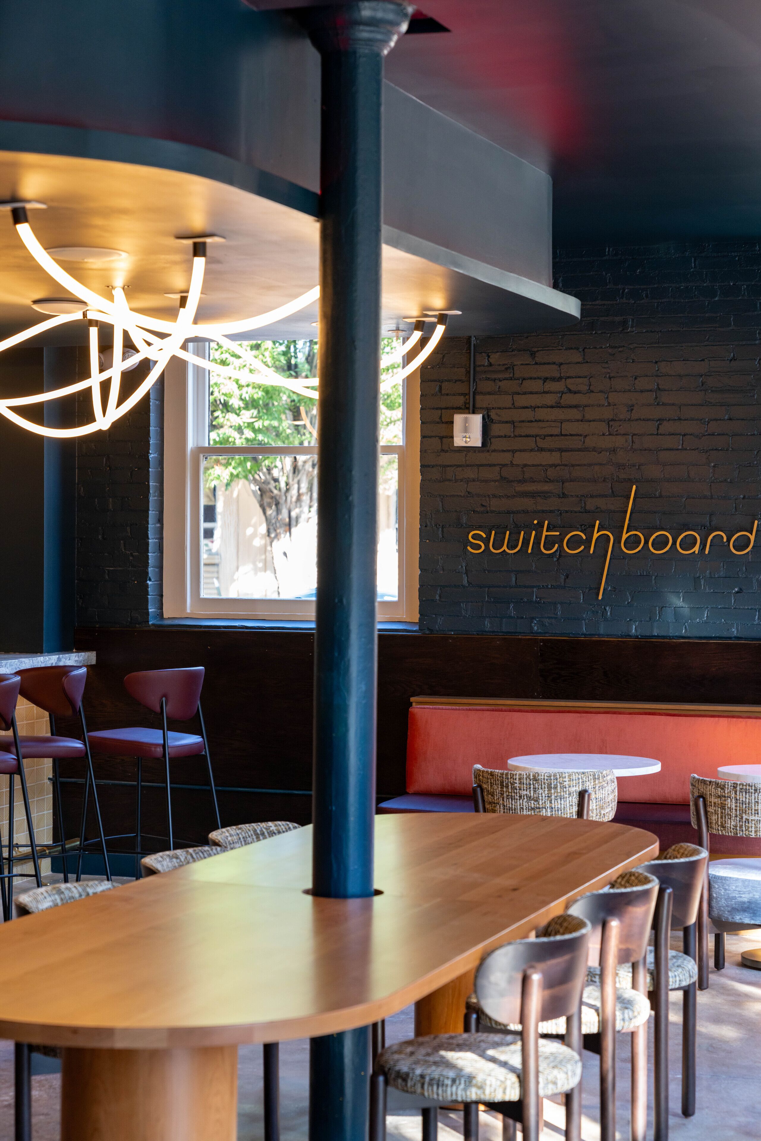 A modern restaurant interior with a long wooden table, wicker chairs, and pendant lighting.