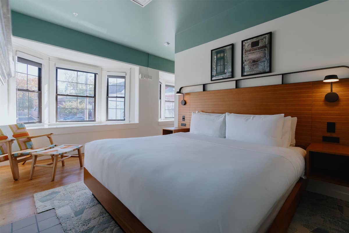 A neatly arranged hotel room with a large bed, wooden headboard, two bedside tables, a chair, and large windows providing natural light.
