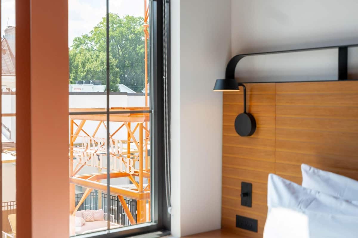 A modern hotel room with a large window, wooden headboard, and a bedside table.