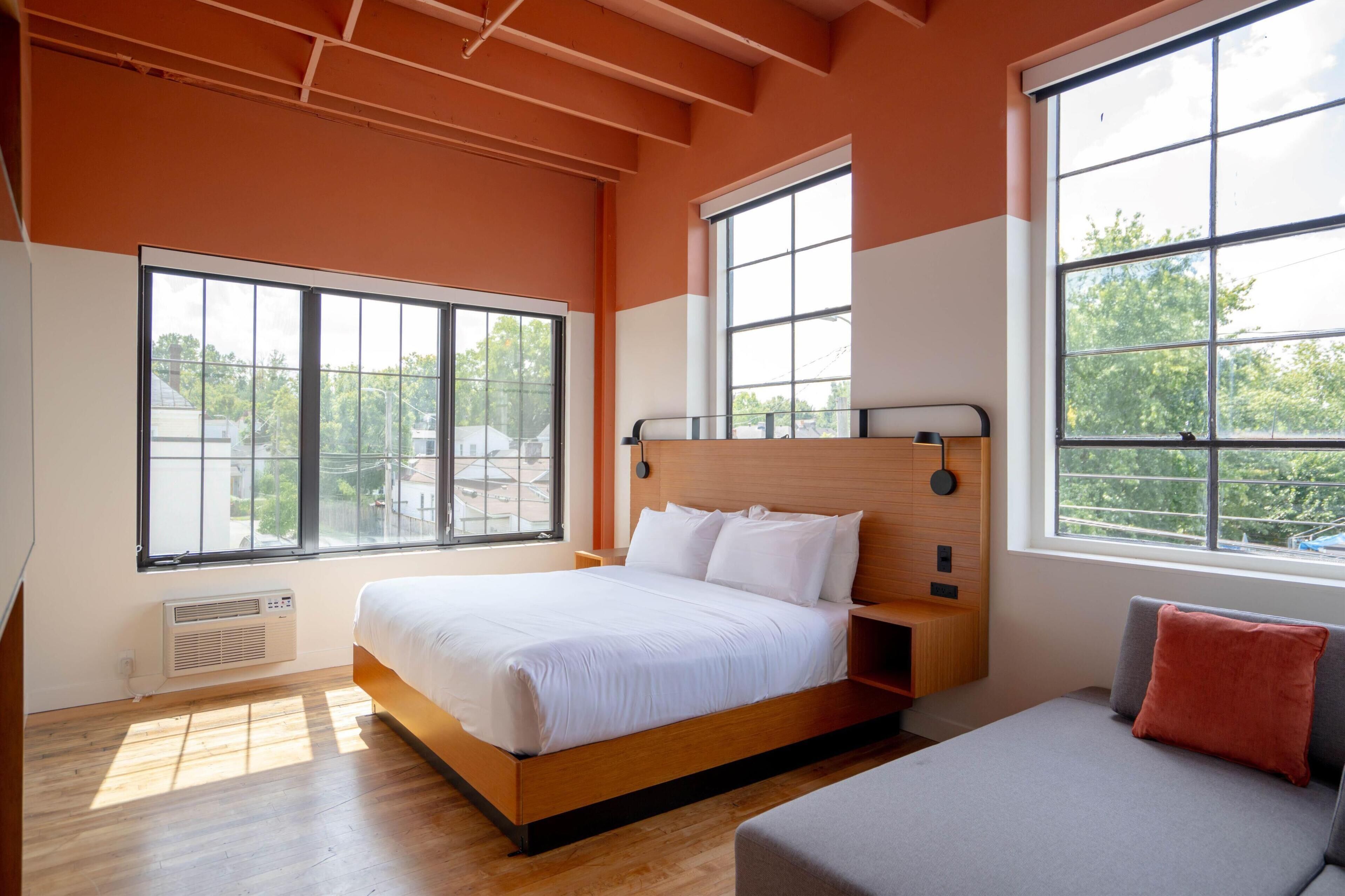 A modern bedroom with a large bed, a sofa, and multiple windows providing natural light.