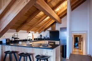 A modern kitchen with wooden beams, a central island with stools, and a view into the living area. - LA JAVA 150 m², rated 5 stars, charm and comfort AT THE FOOT OF THE SLOPES (Villard-Reculas)