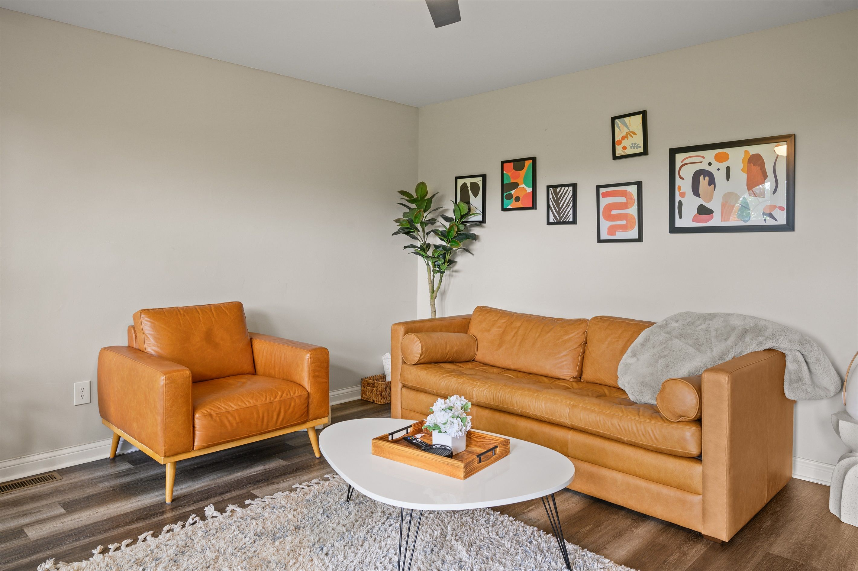 A living room with a brown leather sofa, a white coffee table, and a grey blanket. There are framed artworks on the wall and a potted plant.