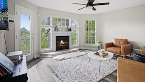Una sala de estar con chimenea, ventanas grandes, un ventilador de techo y un sillón de cuero.