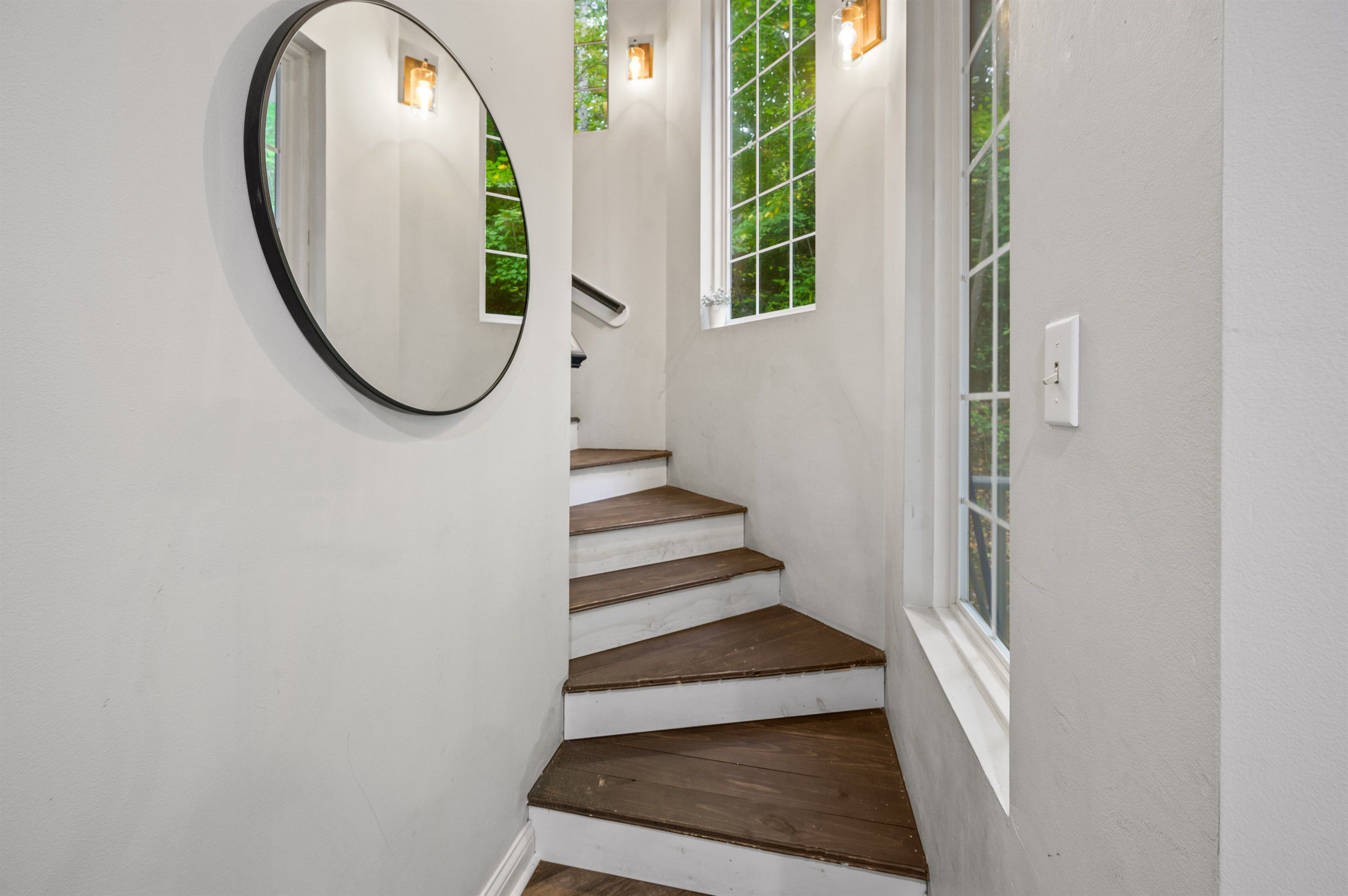 Una escalera con escalones de madera y paredes blancas, un espejo redondo y dos ventanas con luces.