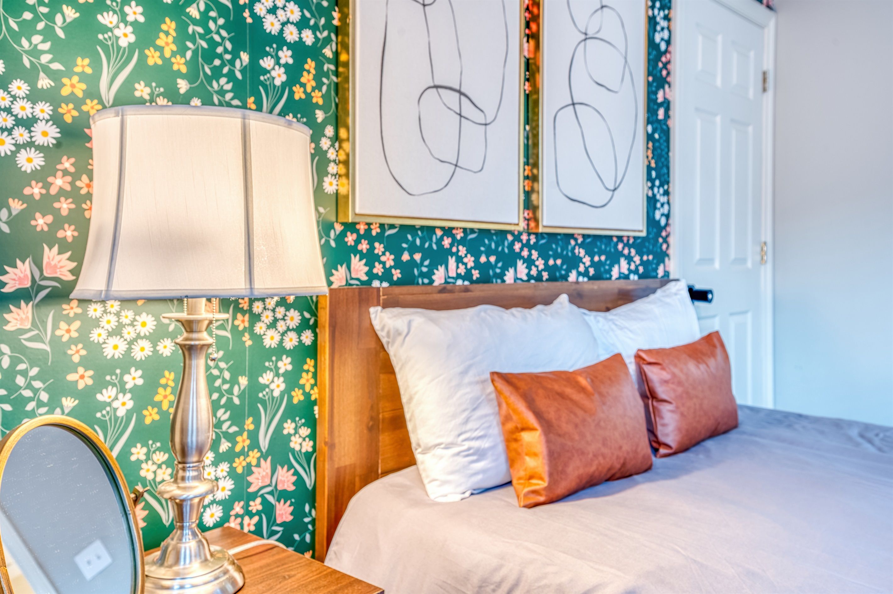 A bedroom with a bed, pillows, a lamp, a mirror, and floral wallpaper.