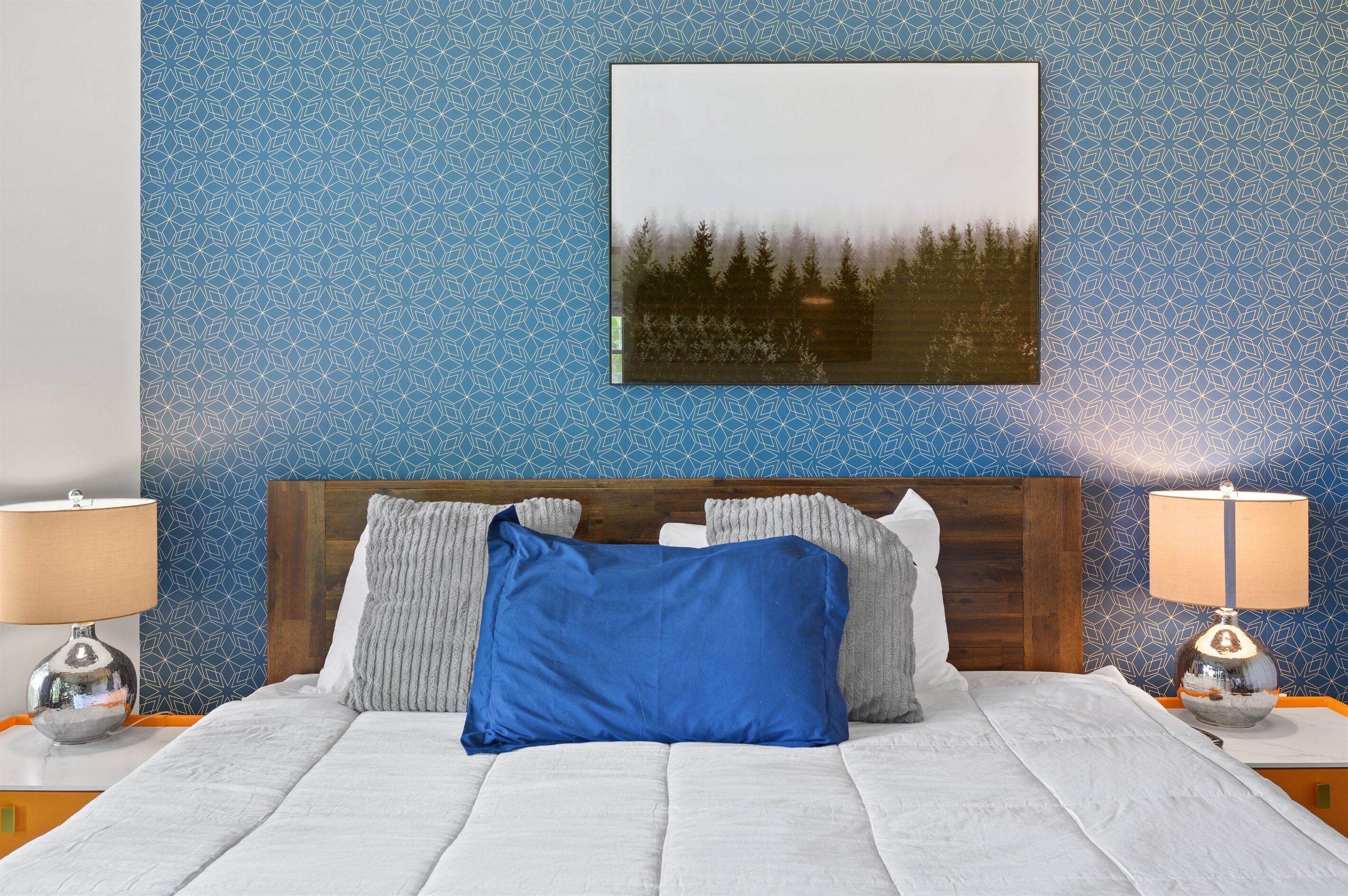 Un lit bien fait, avec un oreiller bleu, une tête de lit en bois et un tableau représentant une forêt accroché au mur.