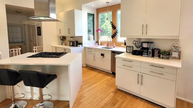 A modern kitchen with white cabinetry, a central island with bar stools, and a view of the outdoors through a window. - Gorgeous Midtown 3 Bedroom Home (Omaha)