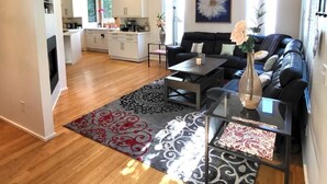 A modern living room with a black leather sofa, a glass coffee table, and a decorative vase. There is a kitchen in the background with white cabinets and a dining area. - Gorgeous Midtown 3 Bedroom Home (Omaha)