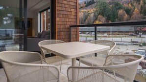 Un balcón con una mesa y sillas, un ventanal de vidrio que da a un cuarto interior, y vistas a un estacionamiento y colinas.