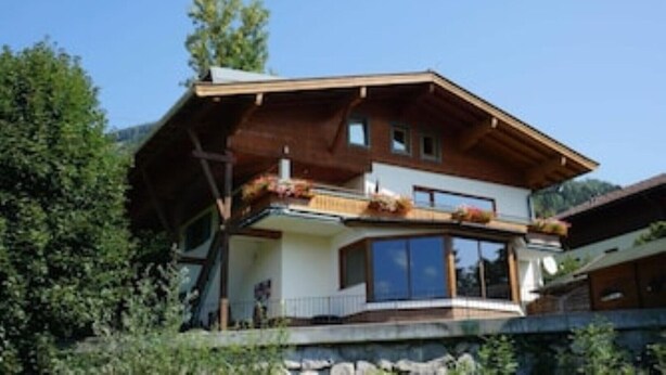 A two-story chalet with a wooden exterior, large windows, and a balcony with plants.