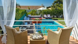 Zona junto a la piscina con mobiliario de mimbre, una mesa con jarras y vasos, y vista a una piscina.