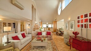 Amplia sala de estar con un sofá blanco, un sillón a rayas y una mesa de centro. Una escalera conduce a un nivel superior, y se observa un comedor al fondo.