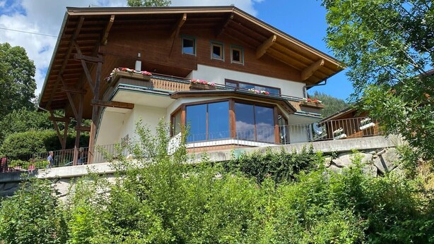 Een chalet met twee verdiepingen, een schuin dak, grote ramen en een balkon met bloembakken.