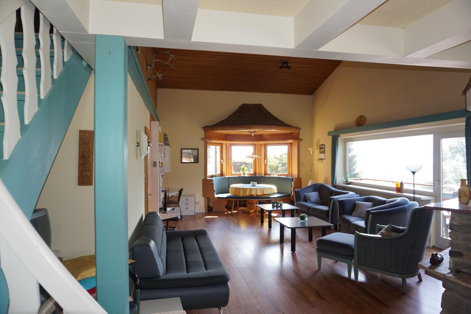 Een ruime woonkamer met een houten plafond, een eethoek met een ronde tafel, en een groot raam dat zorgt voor daglicht.