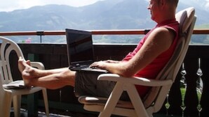 Iemand zit op een stoel met een laptop en kijkt uit over een bergachtig landschap.