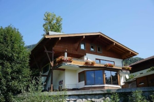A two-story chalet with a wooden exterior, large windows, and a balcony with plants.