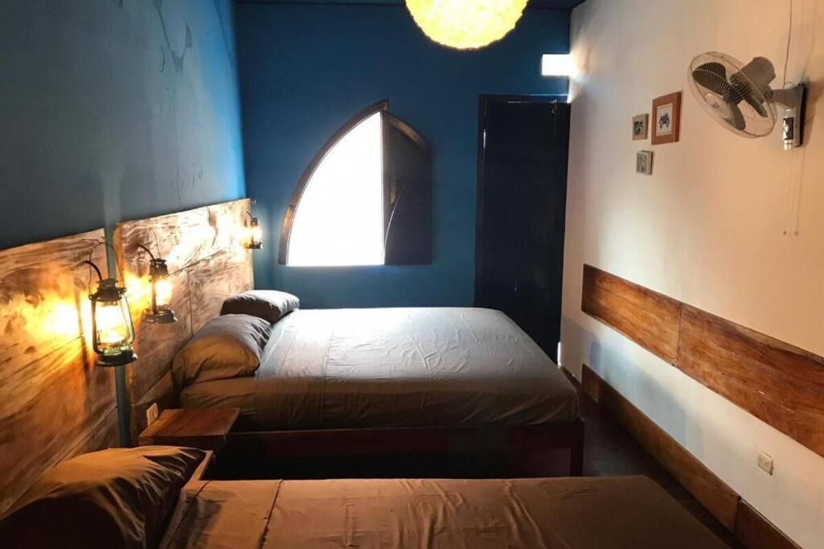 A bedroom with two beds, a wooden headboard, a wall-mounted fan, and a window with natural light.