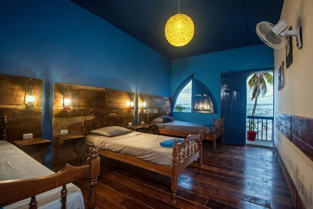 A room with two wooden beds, a hanging light fixture, a fan, and a view of the sea through an arched window.
