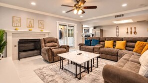 A modern living room with a fireplace, seating area, and a ceiling fan. - Spacious 5BR 2BA Home | Pool, Grill & Game Room (The Colony)
