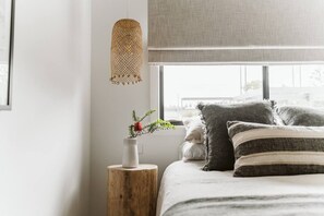 Une chambre à coucher avec un lit, une table de chevet ornée d’un vase de fleurs, un luminaire suspendu et une fenêtre donnant sur des immeubles.